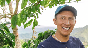 Man wearing a blue cap and shirt standing among green foliage with a scenic background