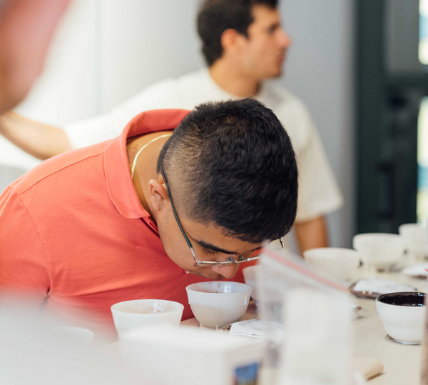 A man in a coral orange polo shirt and glasses leans over a table filled with small white cups, with another man blurred in the background.