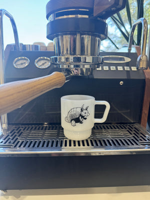 A white ceramic coffee mug with a black armadillo illustration sits on the drip tray of a black espresso machine with a wooden-handled portafilter in the foreground.