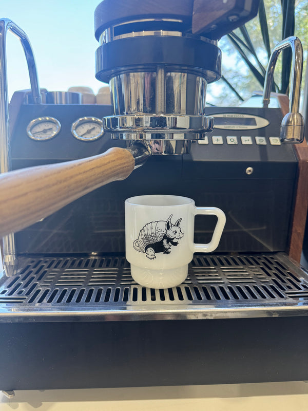 A white ceramic coffee mug with a black armadillo illustration sits on the drip tray of a black espresso machine with a wooden-handled portafilter in the foreground.
