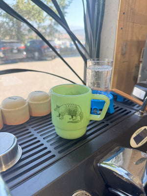 A light green ceramic mug with a black armadillo illustration sits on a black coffee machine drip tray inside what appears to be a camper or RV kitchen. A glass of water and small containers are nearby, with a window and outdoor scene visible in the background.