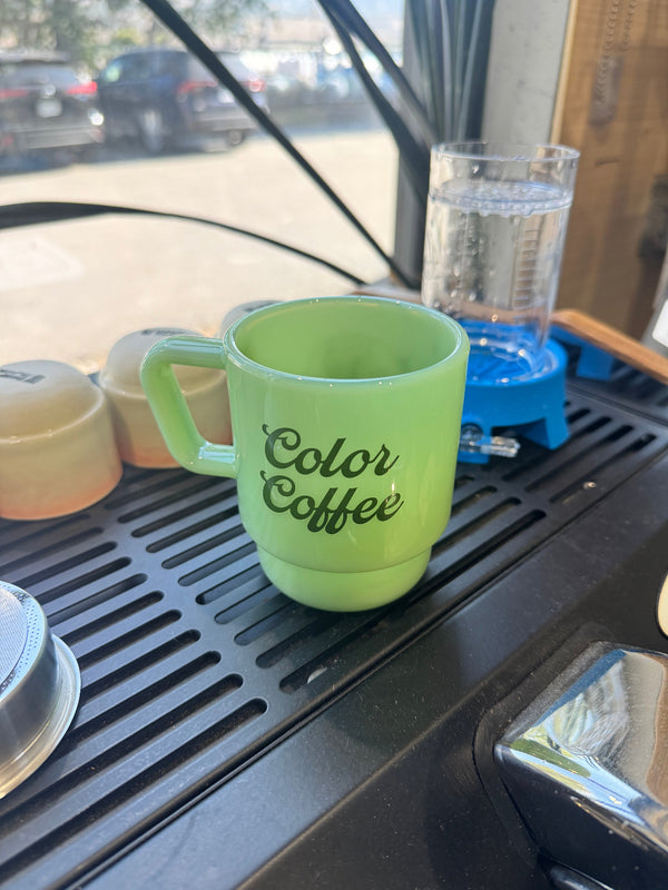 A light green ceramic coffee mug with 'Color Coffee' printed in black script, sitting on a black drip tray inside what appears to be a vehicle or small kitchen setting.