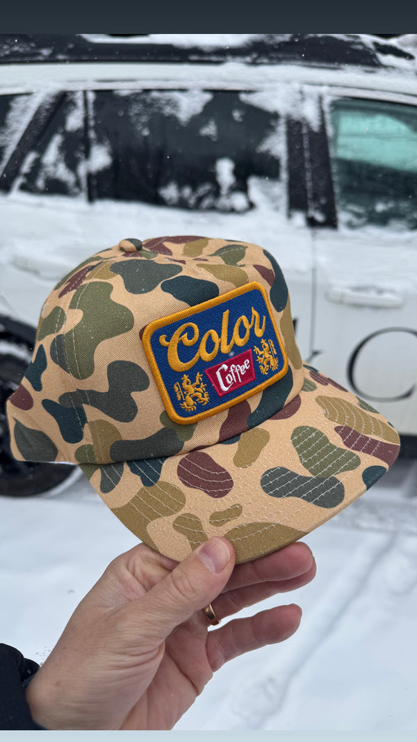 Camouflage cap with a colorful Color patch held in front of a snowy vehicle.