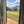 Load image into Gallery viewer, A scenic view of rolling green hills and a grassy field under a blue sky with white clouds, framed by an open rustic wooden door.
