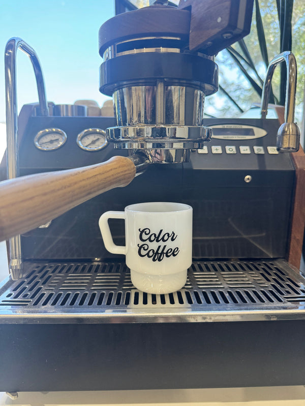 A white ceramic coffee mug with 'Color Coffee' printed in black script sits on the drip tray of a black and wood espresso machine.