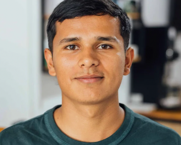 A close-up portrait of a man with dark hair wearing a dark green crewneck t-shirt, set against a softly blurred indoor background.