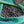 Load image into Gallery viewer, A close-up of dark brown coffee beans spread out in a green mesh sack, with a hand lifting the top edge of the sack.
