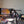 Load image into Gallery viewer, A camera mounted on a tripod set up in front of a large monitor displaying an abstract orange and yellow painting, with various paint bottles, brushes, and a laptop on a desk in a room.
