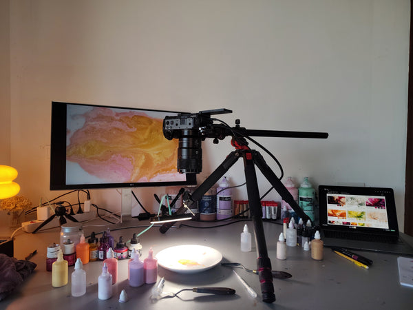 A camera mounted on a tripod set up in front of a large monitor displaying an abstract orange and yellow painting, with various paint bottles, brushes, and a laptop on a desk in a room.