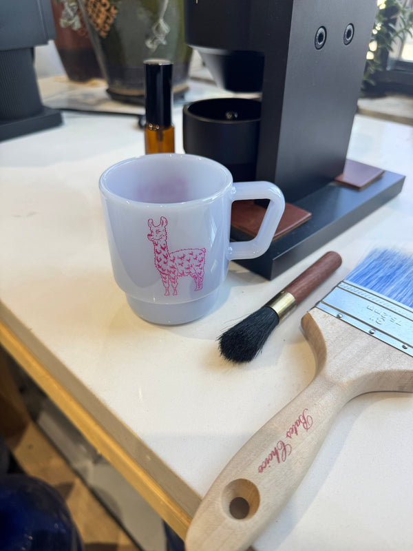 A white ceramic mug with a pink llama pattern sits on a light-colored countertop next to a paintbrush and a paint brush handle, with a coffee machine in the background.